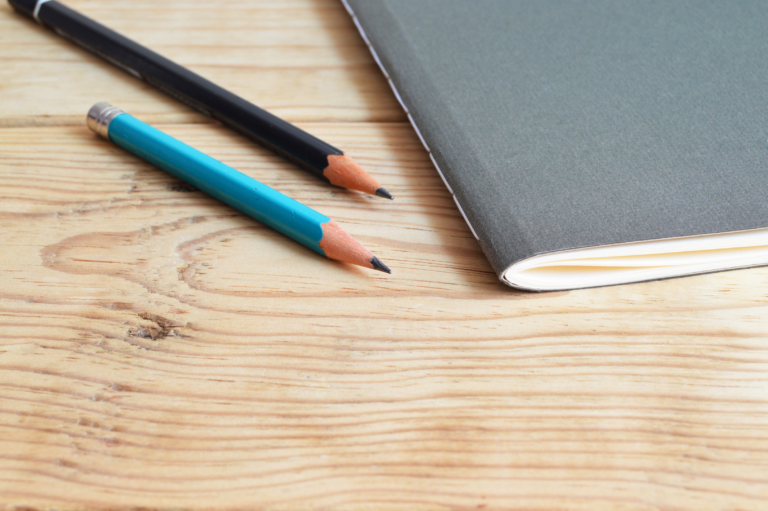 Ein schwarzer und ein blauer Stift neben einem schwarzen Notizheft auf einem Holztisch.