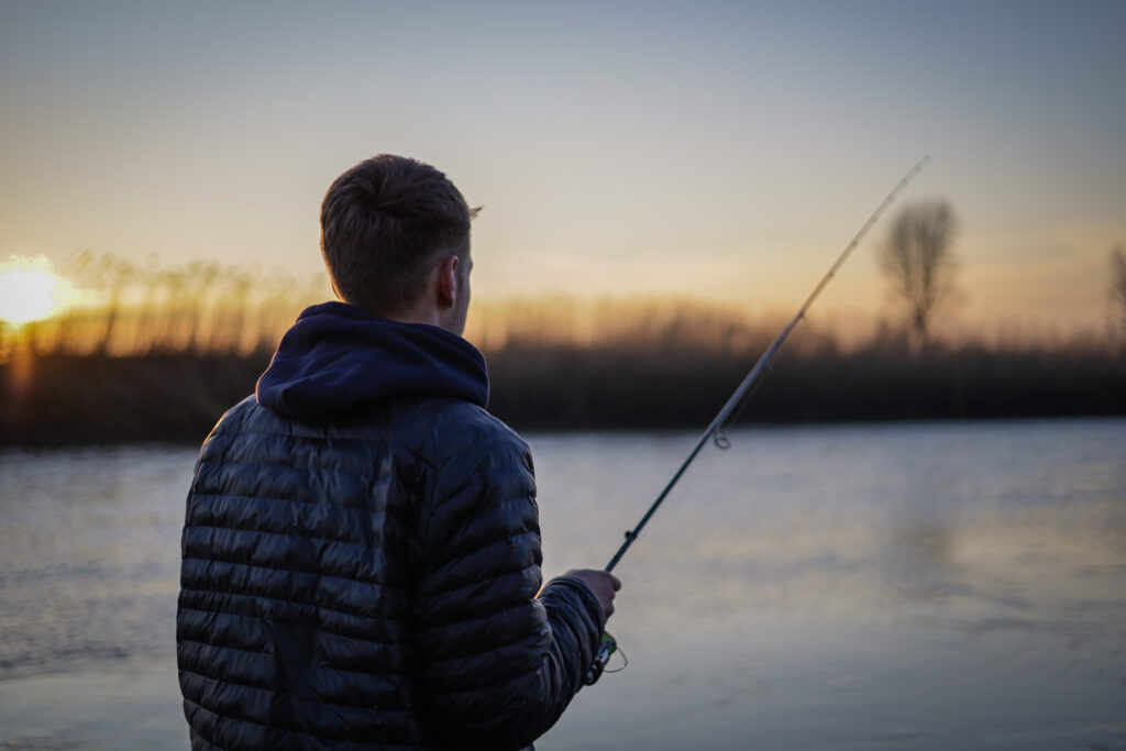 Ein Angler steht an einem Flus und wirft seine Angelrute aus w&auml;hrend im Hintergrund die Sonne untergeht.