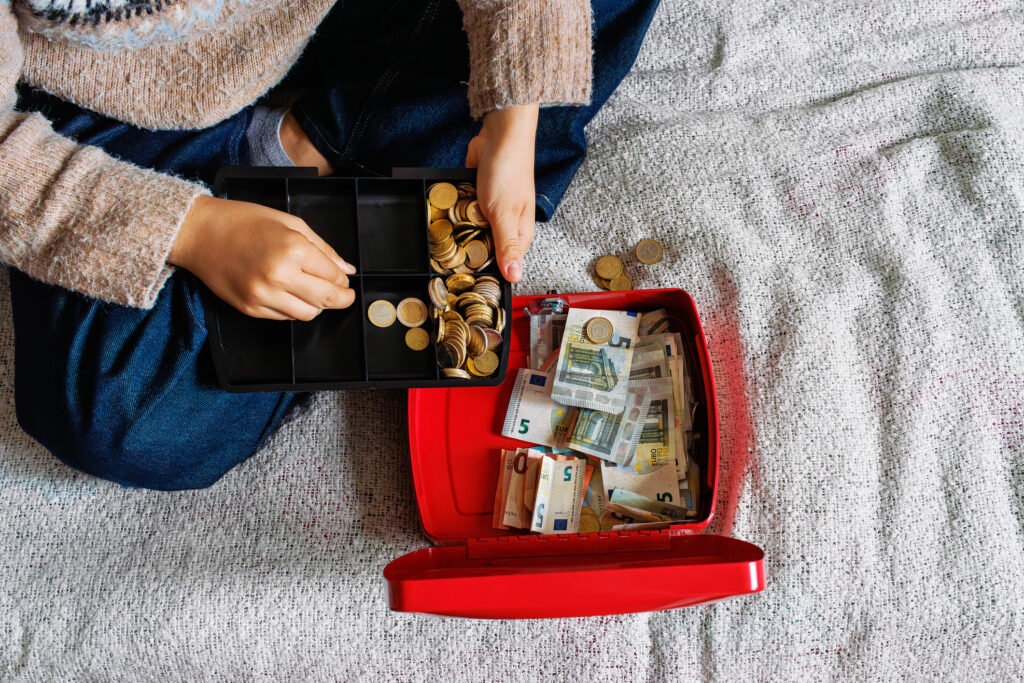 Vogelansicht einer jungen Frau die auf einem Bett sitzt und Eurom&uuml;nzen und Banknoten in einer Geldkasette z&auml;hlt.