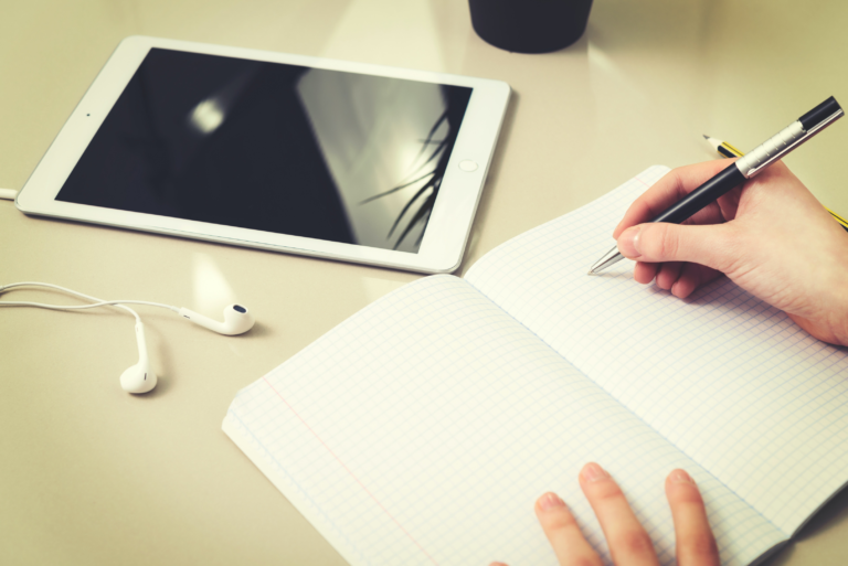 Eine Hand hält einen Kugelschreiber und schreibt in ein leeres Notizbuch, während daneben ein weißes iPad liegt.