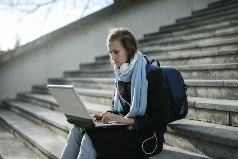 Eine Frau mit blauen Schal sitzt auf einer Betontreppe und arbeitet auf ihrem Laptop auf dem Schoß.