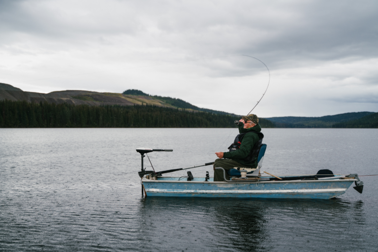Ein Angler sitzt in einem blauen Boot und angelt, wobei im Hintergrund eine Berglandschaft zu sehen ist.