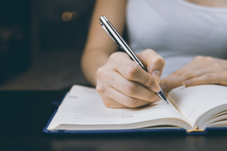 Eine Hand von einer Frau, die in einen Kugelschreiber hält und in das Notizheft schreibt.