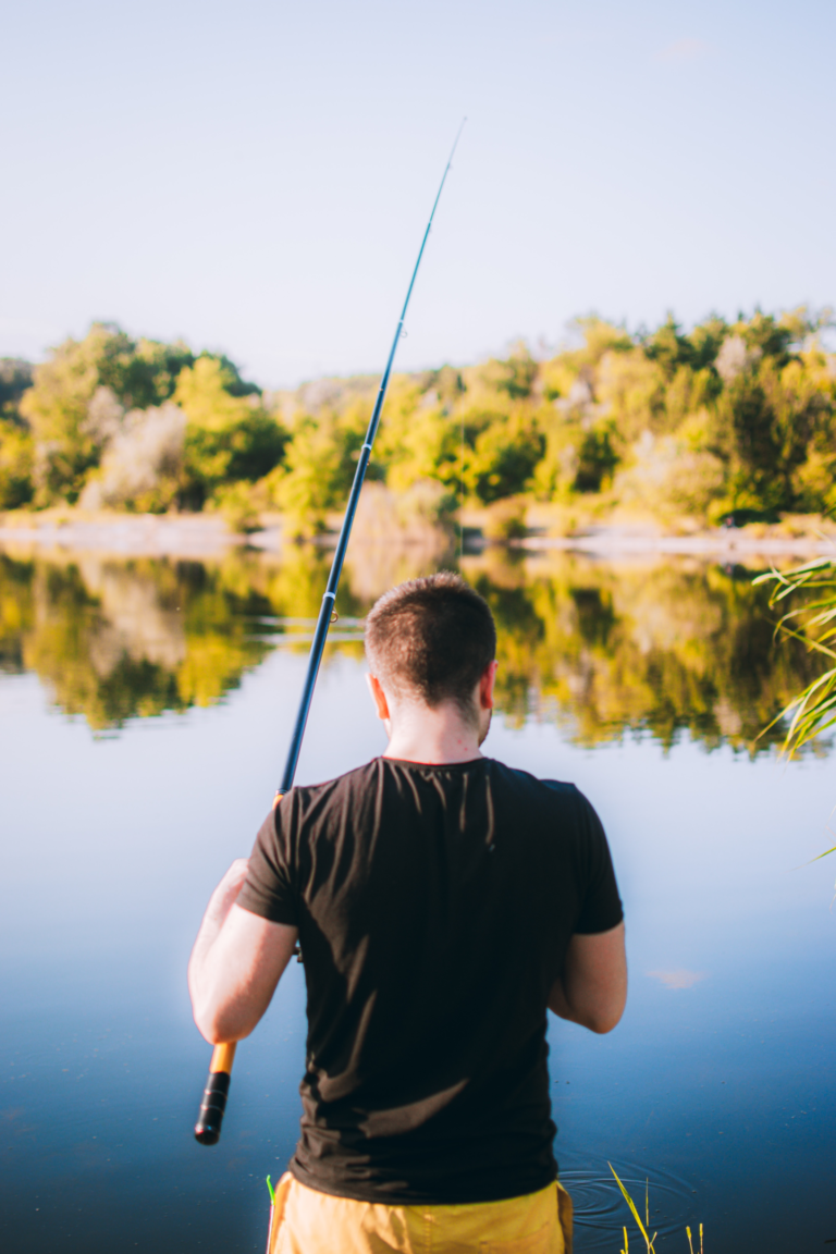 Ein Angler, der am Wasser steht und auf das ruhige Wasser blickt. Im Hintergrund sind grüne Bäume und Büsche zu sehen.