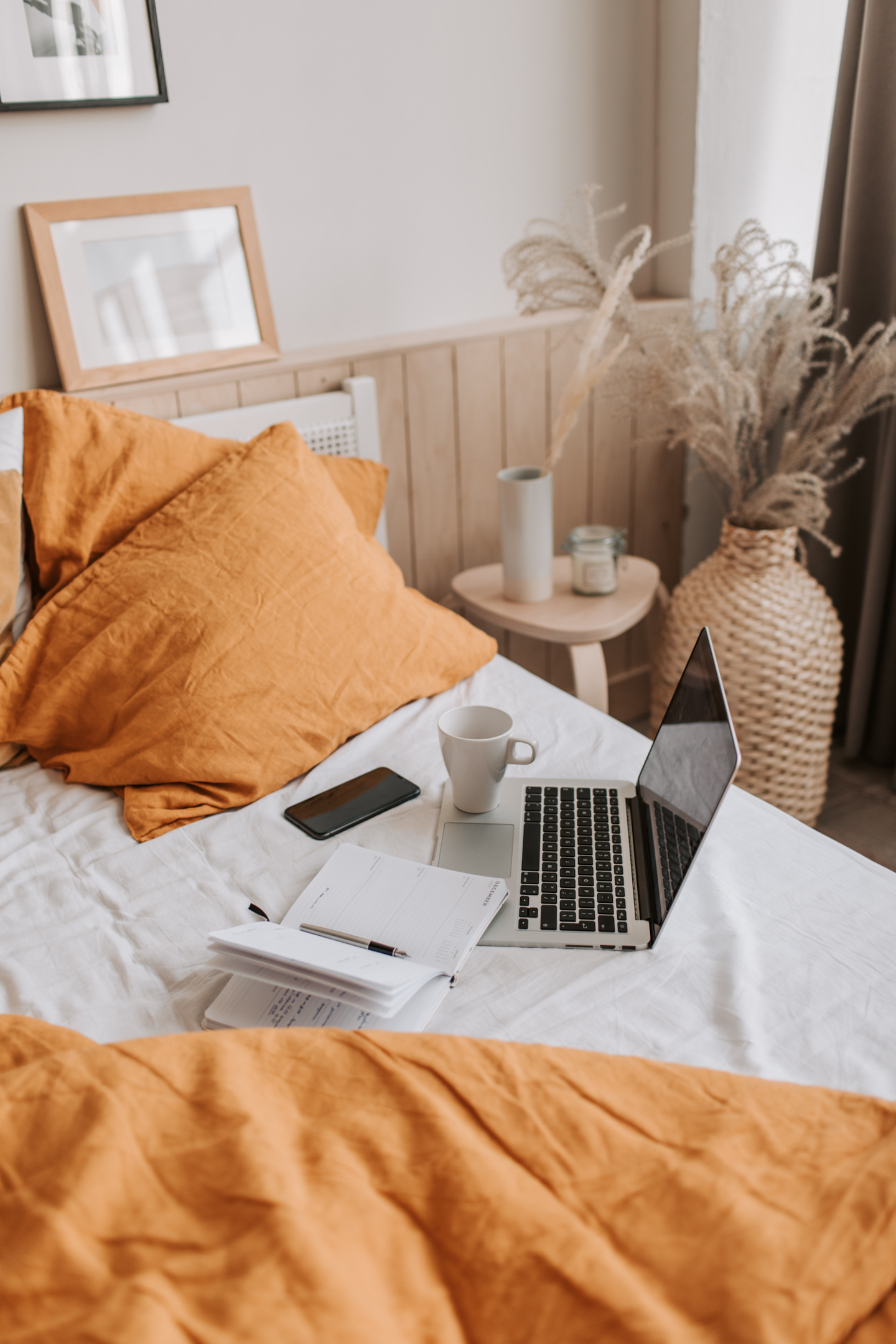 Ein Laptop, ein Handy, eine Tasse sowie ein Notizbuch liegen auf einem Bett mit orangenen Kissen daneben.