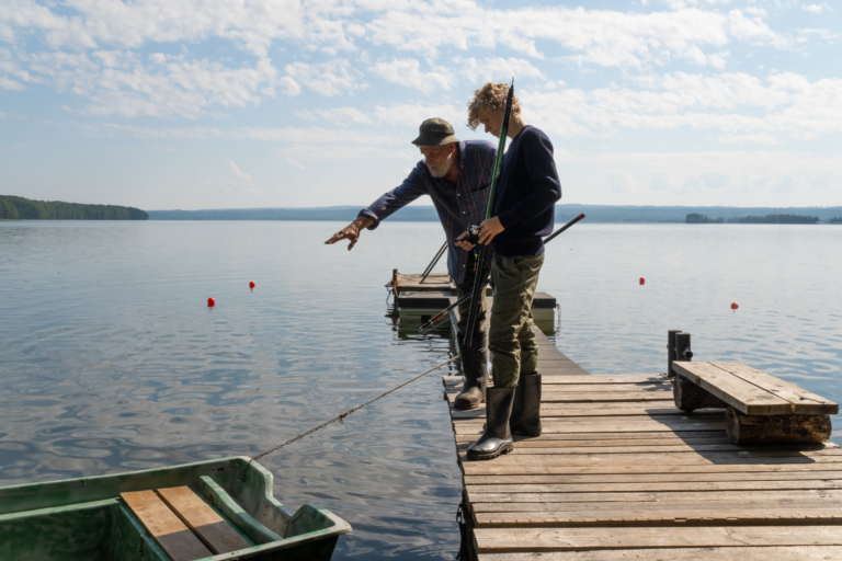 2 Menschen beim Fischen auf einem Holzsteg vor einem Boot in einem See.