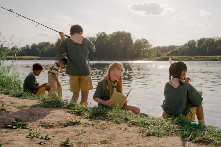 Gruppe von Kindern beim Angeln am See, sie sitzen zusammen am Ufer des Gewässers und halten ihre Angelruten.