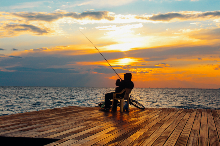 Ein sitzender Angler auf einer Holzplattform am Meer, wobei im Hintergrund die Sonne untergeht.