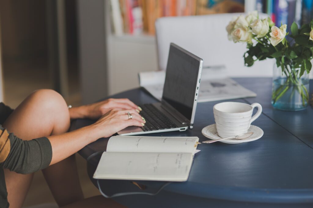 Eine Person, die an einem Tisch sitzt und an einem Laptop arbeitet, wobei daneben eine Tasse und ein Notizbuch sich befinden.