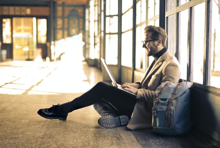 Ein Mann sitzt auf dem Boden und tippt auf einem Laptop, während sein Rucksack neben ihm liegt.