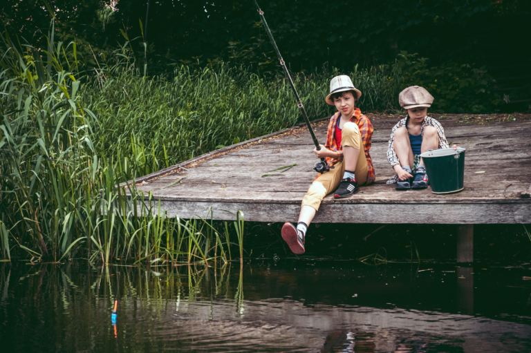 2 Kinder sitzen auf einem Holzsteg und eins hält eine Angelrute. Sie schauen in das Wasser und warten auf einen Biss.